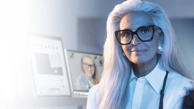 Donna con lunghi capelli grigi e occhiali da vista neri, seduta in un ambiente illuminato da luce blu, con due monitor accesi sullo sfondo. L’immagine comunica un contesto tecnologico e moderno, legato alla protezione visiva dalla luce blu